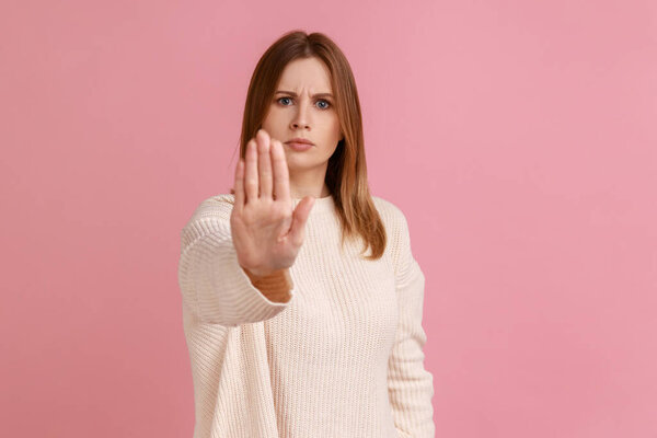 Portrait of serious strict young adult blond woman standing with stop hands gesture and caution to dont touch, wearing white sweater. Indoor studio shot isolated on pink background.