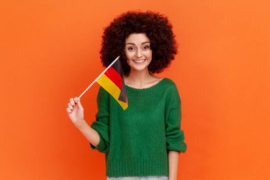 Alman bayrağı tutan, Afro saç stili, yeşil günlük süveter giyen, Alman Birliği Günü 'nü kutlayan çekici, pozitif kadın portresi. Turuncu arka planda kapalı stüdyo çekimleri.