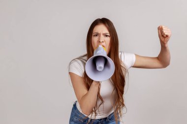 Ağız kenarında megafon tutan, yüksek sesle konuşan, çığlık atan, kolunu kaldırarak anons yapan, protesto eden, beyaz tişört giyen bir kadının portresi. Gri arka planda izole edilmiş kapalı stüdyo görüntüleri.