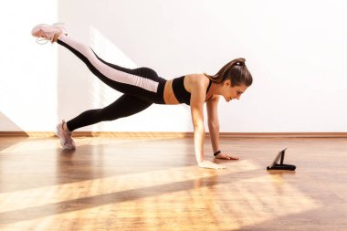 Kalas kaldıran bacaklı bir kadın, tablette özel ders videosu izliyor, spor blogcudan sonra tekrar ediyor, siyah spor bluzu ve tayt giyiyor. Pencereden gelen güneş ışığıyla aydınlatılan tam uzunluktaki stüdyo görüntüleri..