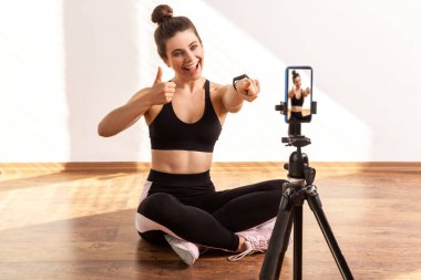 Mutlu fitness eğitmeni telefon kamerasına işaret ediyor ve başparmağını kaldırıyor, online antrenmanından dolayı seni övüyor, siyah spor bluzu ve tayt giyiyor. Pencereden gelen güneş ışığıyla aydınlatılan tam uzunluktaki stüdyo görüntüleri..