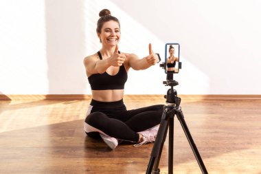 Pozitif fitness koçu telefon kamerasına baş parmak gösteriyor, takipçilerle iyi bir online antrenman yapıyor, siyah spor bluzu ve tayt giyiyor. Pencereden gelen güneş ışığıyla aydınlatılan tam uzunluktaki stüdyo görüntüleri..