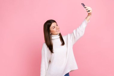 Esmer bir kadın selfie çekiyor ya da canlı yayın yapıyor, cep telefonu kamerasına gülümsüyor, beyaz bir süveter giyiyor. Pembe arka planda kapalı stüdyo çekimleri.