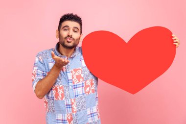 Büyük kırmızı kalbi olan, kameraya bakan, hava öpücüğü gönderen, mavi bir gömlek giyen mutlu, romantik sakallı bir adamın portresi. Pembe arka planda kapalı stüdyo çekimleri.