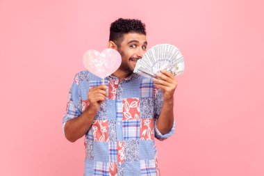 Sakallı mutlu bir adamın portresi dişlek gülüşlü kameraya bakıyor, kağıt kalp ve dolar banknotları tutuyor, mavi gömlek giyiyor. Pembe arka planda kapalı stüdyo çekimleri.