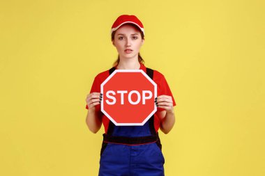 Ciddi işçi kadın portresi kırmızı dur işareti gösteriyor, kameraya kendine güvenen katı bir ifadeyle bakıyor, tulum giyiyor ve kırmızı şapka takıyor. Sarı arka planda kapalı stüdyo çekimleri.