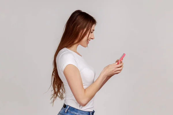 携帯電話を使用して幸せな女性の側のビューは 楽しそうに笑みを浮かべて 入力したり ソーシャルネットワーク上で面白いメッセージを読んで 白いTシャツを着て 灰色の背景に隔離された屋内スタジオショット — ストック写真