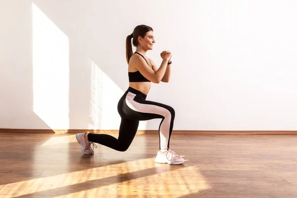 Slim female doing sport lunge exercise, standing one knee, warms up training muscles, butts workout, wearing black sports top and tights. Full length studio shot illuminated by sunlight from window.