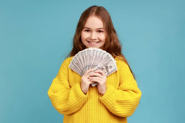 小さな女の子ドル紙幣のファンを保持 大きなお金を示す おもちゃの笑顔でカメラを見て 黄色のカジュアルなスタイルのセーターを着て 青い背景に隔離された屋内スタジオショット — ストック写真