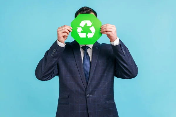 公式スタイルのスーツを身に着けている未知の男性は緑のリサイクル記号 ゴミの分類と環境保護の後ろに顔を隠し 緑を考えます 青い背景に隔離された屋内スタジオショット — ストック写真
