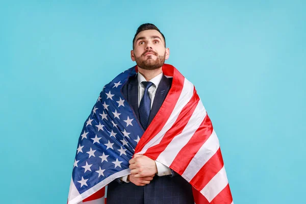 Young adult bearded businessman being wrapped in USA flag, celebrating labor day or US Independence day 4th of july, government employment support. Indoor studio shot isolated on blue background.