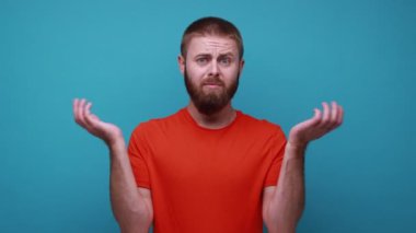 Bilmiyorum, kimin umrunda. Kafası karışık, sakallı, omuzlarını silken, jest yapan, turuncu tişört giyen, kafası karışık bir adam. Mavi arka planda kapalı stüdyo çekimi.
