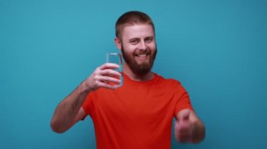 Olumlu iyimser sakallı bir adam elinde bir bardak temiz su tutuyor ve kameraya baş parmağını gösteriyor, içki içmeye çağırıyor, turuncu tişört giyiyor. Mavi arka planda kapalı stüdyo çekimi.