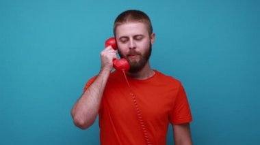 Saldırgan sakallı adam bağırıyor ve eski telefon hattından bağırıyor. Bağlantı kalitesinden şikayet ediyor. Turuncu tişört giyiyor. Mavi arka planda kapalı stüdyo çekimi.