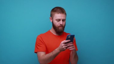 Şaşırmış genç sakallı adam sosyal ağda şok edici bir yazı okuyor cep telefonu kullanıyor, şaşırmış görünüyor, turuncu tişört giyiyor. Mavi arka planda kapalı stüdyo çekimi.