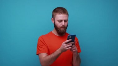 Akıllı telefonu elinde tutan dalgın sakallı adam, yazılım güncellemesi hakkında kafa yoruyor, mesajlara ne cevap vereceğini düşünüyor, turuncu tişört giyiyor. Mavi arka planda kapalı stüdyo çekimi.