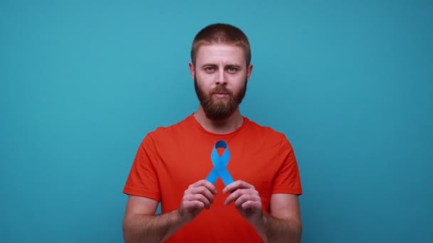 积极乐观的大胡子男人拿着蓝丝带 看着相机 露出大拇指 医学概念 穿着橙色T恤 室内拍摄被蓝色背景隔离 — 图库视频影像