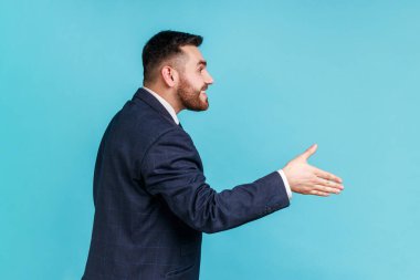 Merhaba, tanıştığımıza memnun oldum! Resmi tarz takım elbise giymiş, iş görüşmesinde el sıkışmayı öneren güler yüzlü yakışıklı bir adamın yan görüntüsü. Mavi arka planda kapalı stüdyo çekimi.