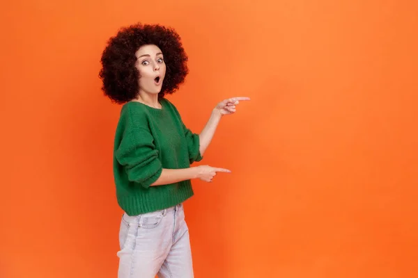 顔の表情を驚かせた緑のセーターの脇に指を指してアフロの髪型を持つ驚くべき女性は 口を開いて コピースペースを維持します オレンジの背景に隔離された屋内スタジオショット — ストック写真