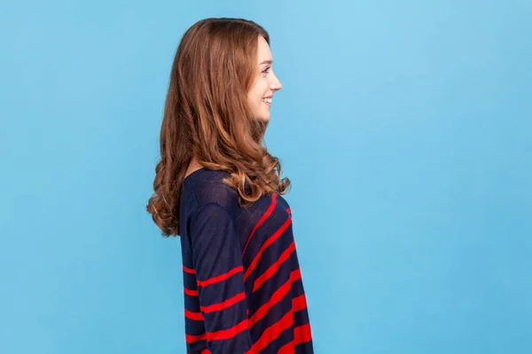 縞模様のカジュアルなスタイルのセーターを着て笑顔の女性の側のビューに立って先を見て 肯定的な表現をして 良い気分にされます 青い背景に隔離された屋内スタジオショット — ストック写真