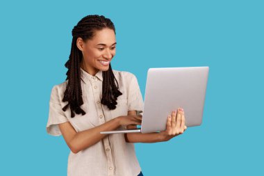 Elinde dizüstü bilgisayarla gülümseyen kadın, sosyal ağlardan komik videolar izlerken gülüyor, internette sörf yapıyor, beyaz gömlek giyiyor. Mavi arka planda kapalı stüdyo çekimi.