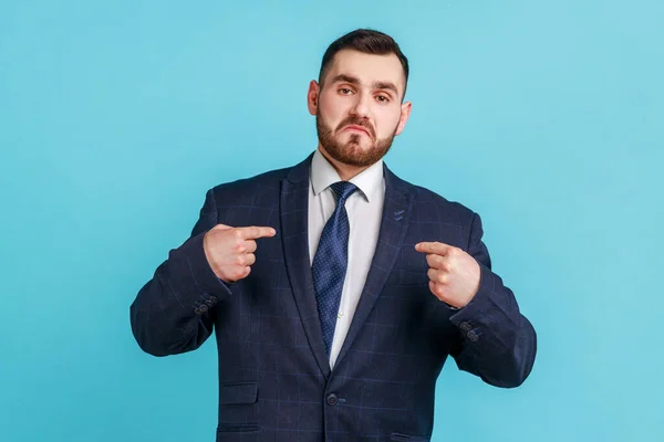This is me! Portrait of egoistic arrogant selfish man with beard wearing official style suit pointing himself, boasting successful achievement. Indoor studio shot isolated on blue background.
