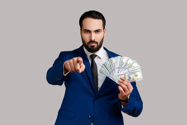 严肃而专横的大胡子男人拿着美元钞票指着相机 主动提供赚钱的机会 穿着正式风格的西服 在灰色背景下被隔离的室内拍摄 — 图库照片