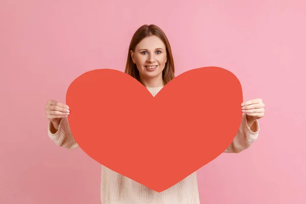 幸せなロマンチックなブロンドの女性の肖像カメラに大きな赤いハートを保持し 愛と肯定的な感情を表現し 白いセーターを着ている ピンクの背景に隔離された屋内スタジオショット — ストック写真
