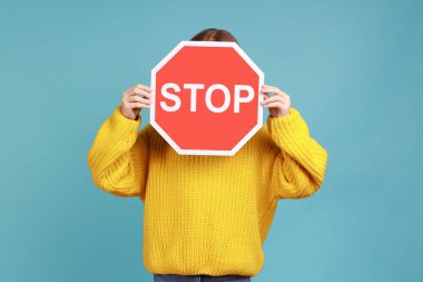 Bilinmeyen küçük çocuğun portresi yüzünü dur sembolüyle kaplıyor, kimliği belirsiz çocuk kırmızı trafik işareti tutuyor, sarı günlük süveter giyiyor. Mavi arka planda kapalı stüdyo çekimi.