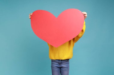 Büyük kırmızı kalbin arkasına saklanan küçük bir kızın portresi, aşk ve sevginin sembolü, sarı günlük süveter giyiyor. Mavi arka planda kapalı stüdyo çekimi.