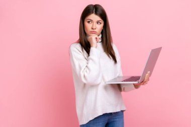 Düşünceli bir kadının portresi dizüstü bilgisayarını tutuyor, elini çenesine koyuyor, yeni bir proje düşünüyor, günlük beyaz süveter giyiyor. Pembe arka planda kapalı stüdyo çekimleri.