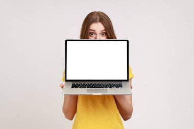 Yüzünün yarısını laptopun arkasına saklayan sarı tişörtlü, beyaz boş ekranlı, büyük şaşırmış gözlerle kameraya bakan kimliği belirsiz bir kadın var. Gri arka planda izole edilmiş kapalı stüdyo görüntüleri.