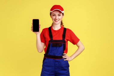 Genç ve çekici bir kadın elinde akıllı bir telefonla reklam için boş bir ekran gösteriyor, tulum ve kırmızı başlık takıyor. Sarı arka planda kapalı stüdyo çekimleri.