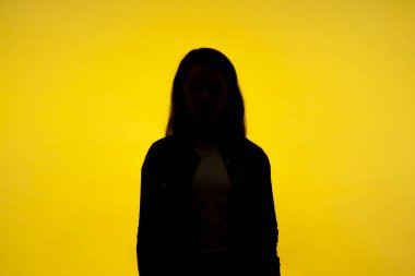 İsim yok, yüzünü gölgede saklayan isimsiz biri, insan kimliği. Kimliği belirsiz bir kadının siluet portresi. Tek başına ayakta duruyor. Stüdyo resmi, sarı arka planda izole edilmiş.