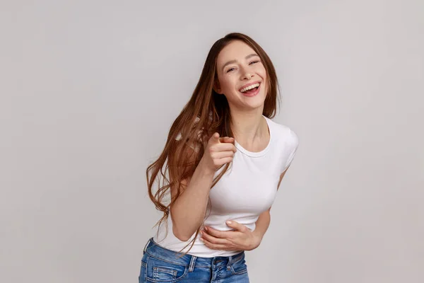 Hey, you are ridiculous. Joyful woman laughing, holding stomach, pointing to camera, taunting you, can\'t stop hysterical laughter, wearing white T-shirt. Indoor studio shot isolated on gray background