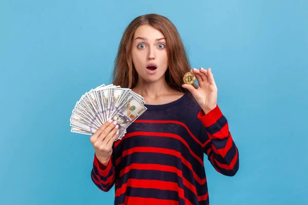 縞模様のカジュアルなスタイルのセーターを着て口を開けて驚くべき女性は ドル紙幣やビットコイン デジタルマネー 電子商取引を示しています 青い背景に隔離された屋内スタジオショット — ストック写真