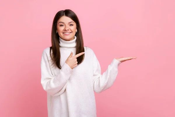 白いカジュアルなスタイルのセーターを着て 商業的なテキストや商品のための空の場所を示す 彼女の手のひらにコピースペースを提示幸せな女性 ピンクの背景に隔離された屋内スタジオショット — ストック写真