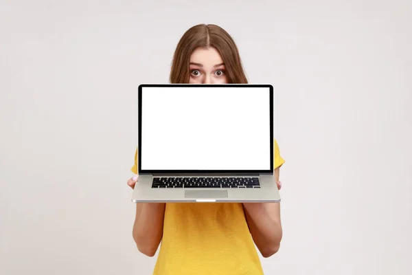 黄色のTシャツに肯定的な未知の女性の白い空のディスプレイとノートパソコンの後ろに顔の半分を隠し 大きな驚きの目でカメラを見て 灰色の背景に隔離された屋内スタジオショット — ストック写真