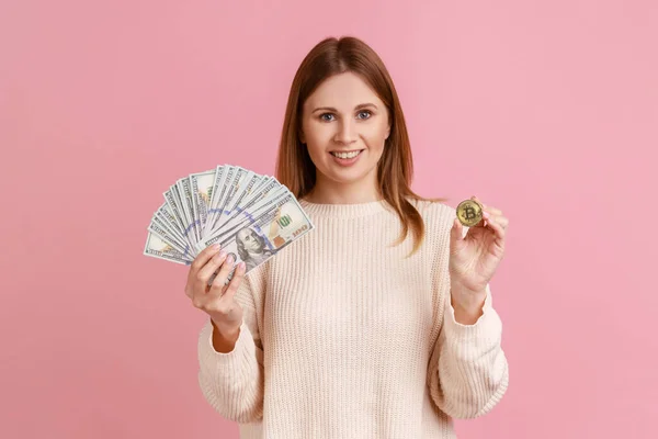 手に握って 白いセーターを着て デジタルビットコインにお金を転送し 暗号通貨のドル紙幣と黄金のコインを示す女性 ピンクの背景に隔離された屋内スタジオショット — ストック写真