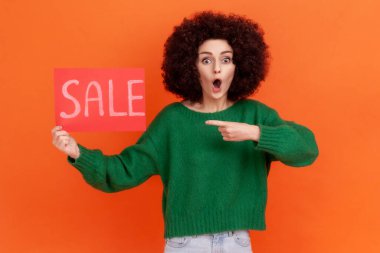 Yeşil kazaklı, Afro saç stili genç bir kadın. Üzerinde satılık bir kart olan kazağı gösteriyor. Şok olmuş bir ifadeyle kameraya bakıyor. Turuncu arka planda kapalı stüdyo çekimleri.