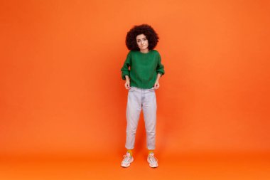 Afro saç stili, yeşil günlük süveter giyen, boş cepleri olan, işsiz bir kadının tam boy portresi. Turuncu arka planda kapalı stüdyo çekimleri.