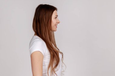 Çekici gülüşü olan güzel bir kadının yan görünüş portresi. Durup geleceğe bakıyor, olumlu duygularını ifade ediyor, beyaz tişört giyiyor. Gri arka planda izole edilmiş kapalı stüdyo görüntüleri.