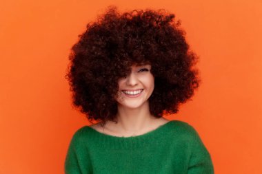 Afro saç stili gülümseyen kadın yeşil günlük bir süveter giyiyor kameraya bakıyor, kabarık saçlarını gösteriyor, güzellik salonunun reklamını yapıyor. Turuncu arka planda kapalı stüdyo çekimleri.