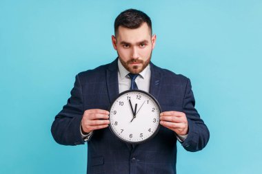 Elinde resmi bir takım elbiseyle patronluk taslayan ciddi bir adam. Büyük duvar saati, canı sıkkın bir şekilde kameraya bakıyor. Harekete geçme zamanı, geri sayım. Mavi arka planda kapalı stüdyo çekimi.