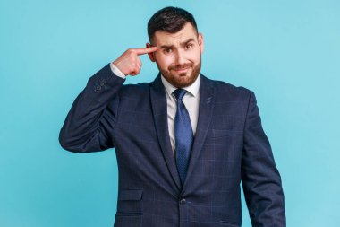 Crazy idea! Portrait of man in dark suit showing stupid gesture, looking at camera with condemnation and blaming for insane plan, dumb suggestion. Indoor studio shot isolated on blue background.