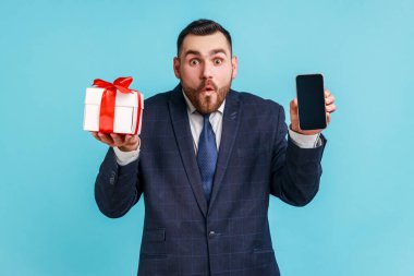 Vay canına, seyyar kullanıcı için hediye ikramiyesi! Koyu renk takım elbiseli, cep telefonlu ve reklam için boş pozlar veren genç ve hayrete düşmüş bir adam. Mavi arka planda kapalı stüdyo çekimi.