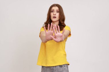 Kahverengi dalgalı saçlı, hoşnutsuz bir kadının portresi. İki eliyle de dur işareti yapıyor. Günlük sarı tişört giyiyor. Gri arka planda izole edilmiş kapalı stüdyo görüntüleri.