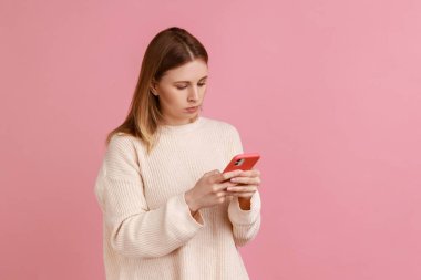 Cep telefonu kullanan, cihaz ekranına bakan, beyaz kazak giyen ciddi bir şekilde konsantre olmuş sarışın kadının portresi. Pembe arka planda kapalı stüdyo çekimleri.