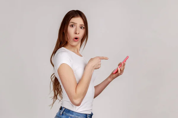 驚いた女性は 携帯電話を使用して チャット スマートフォンの画面を指して 白いTシャツを着て ソーシャルネットワーク上でショックを受けた投稿を読んで 灰色の背景に隔離された屋内スタジオショット — ストック写真