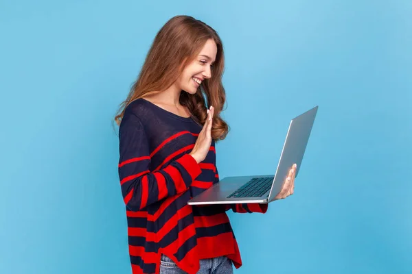 縞模様のカジュアルなスタイルのセーターを着て陽気な女性は ラップトップで立って ディスプレイを見て手を振って こんにちは ビデオ通話を持っていると言います — ストック写真